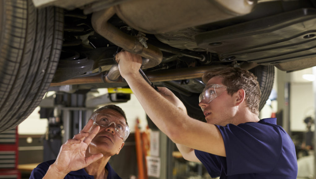 Mechanic And Male Trainee Working Underneath Car Together