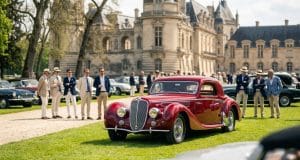 Concours d’élégance automobile : l’art de vivre français Une élégante voiture de sport rouge classique exposée devant un château français avec des hommes en costumes et chapeaux.