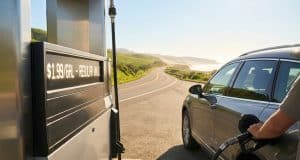 Prix du carburant cet été : prévisions pour vos vacances Gros plan sur une main faisant le plein d'une voiture dans une station-service avec une route côtière en arrière-plan.