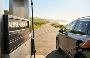 Prix du carburant cet été : prévisions pour vos vacances Gros plan sur une main faisant le plein d'une voiture dans une station-service avec une route côtière en arrière-plan.