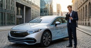 Xiaomi SU7 en Europe : les risques de l’importation grise Un homme d'affaires pensif examine une tablette devant une Xiaomi SU7 argentée garée sur des pavés en ville.