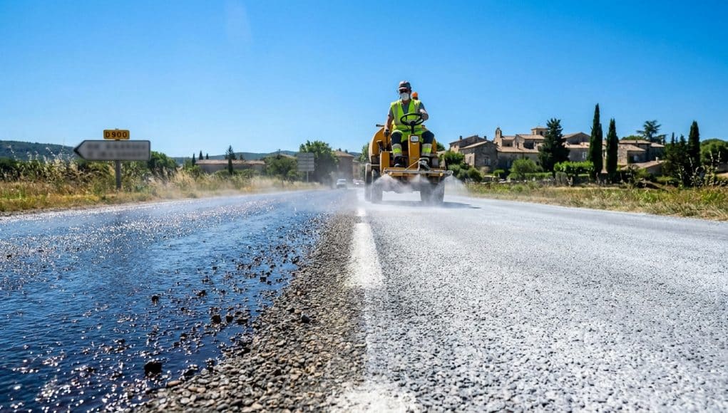 Un ouvrier en engin pulvérise du lait de chaux blanc sur une route sombre pour réduire la chaleur lors d'une canicule.