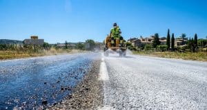 Lait de chaux et routes : la solution face à la canicule Un ouvrier en engin pulvérise du lait de chaux blanc sur une route sombre pour réduire la chaleur lors d'une canicule.