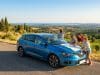 Les vacances en voiture : pilier de l’été français en 2026 Une famille regarde une carte sur le capot d'une voiture bleue garée au bord d'une route surplombant des vignobles.