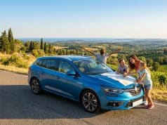Les vacances en voiture : pilier de l’été français en 2026 Une famille regarde une carte sur le capot d'une voiture bleue garée au bord d'une route surplombant des vignobles.
