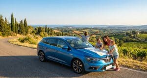 Les vacances en voiture : pilier de l’été français en 2026 Une famille regarde une carte sur le capot d'une voiture bleue garée au bord d'une route surplombant des vignobles.