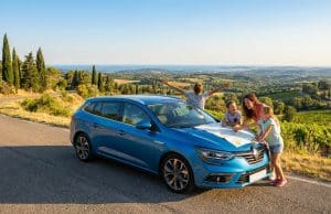 Les vacances en voiture : pilier de l’été français en 2026 Une famille regarde une carte sur le capot d'une voiture bleue garée au bord d'une route surplombant des vignobles.