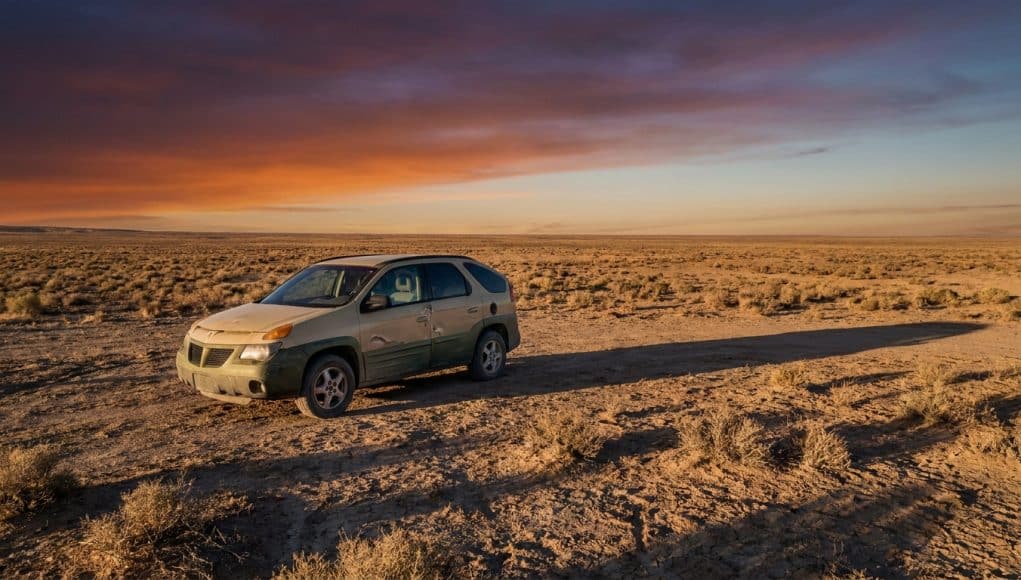 Une Pontiac Aztek beige stationnée seule dans un désert aride sous un coucher de soleil orange et violet.