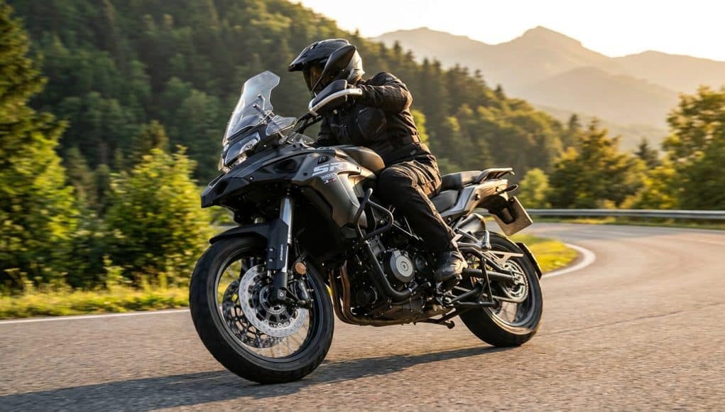 Essai de la moto trail QJ Motor SRT 450 RX Un motard en équipement noir chevauche une moto trail QJ Motor SRT 450 RX grise sur une route sinueuse en montagne.