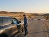 Zeekr bloqué à la frontière : le contrôle logiciel en débat Un homme debout devant une voiture Zeekr bloquée par une barrière sur une route isolée dans un paysage désertique.