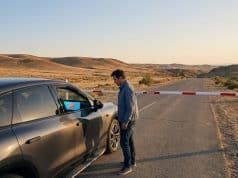 Zeekr bloqué à la frontière : le contrôle logiciel en débat Un homme debout devant une voiture Zeekr bloquée par une barrière sur une route isolée dans un paysage désertique.
