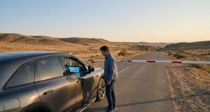 Zeekr bloqué à la frontière : le contrôle logiciel en débat Un homme debout devant une voiture Zeekr bloquée par une barrière sur une route isolée dans un paysage désertique.