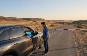 Zeekr bloqué à la frontière : le contrôle logiciel en débat Un homme debout devant une voiture Zeekr bloquée par une barrière sur une route isolée dans un paysage désertique.