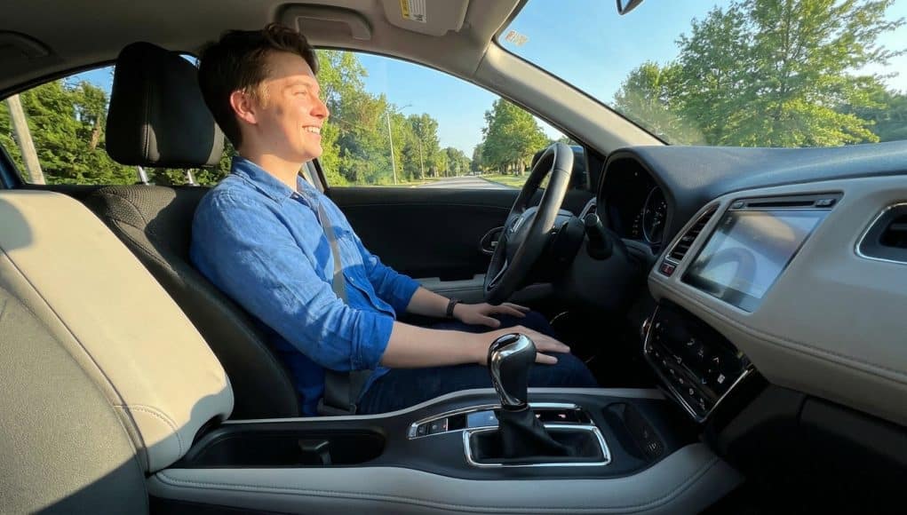 Apprendre à conduire en boîte automatique Un jeune homme souriant au volant d'une voiture à boîte automatique lors d'une journée ensoleillée.