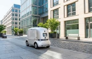 Robot de livraison autonome en France : le nouveau cadre 2026 Un petit robot de livraison blanc autonome circule sur une rue piétonne pavée bordée d'immeubles de bureaux modernes.