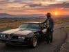 Burt Reynolds et sa Pontiac Trans Am : un accord de légende Un homme en chapeau de cowboy accoudé à une Pontiac Trans Am noire sur une route déserte au coucher du soleil.