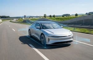 Tesla FSD en France : l’UTAC teste le système de Tesla Une berline électrique argentée roule sur autoroute, entourée de graphiques holographiques illustrant la conduite autonome.