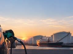 Le prix du gazole baisse de 9 centimes par litre en Europe Pistolet à essence inséré dans une voiture avec en arrière-plan un pétrolier et des réservoirs au coucher du soleil.