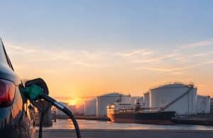 Le prix du gazole baisse de 9 centimes par litre en Europe Pistolet à essence inséré dans une voiture avec en arrière-plan un pétrolier et des réservoirs au coucher du soleil.