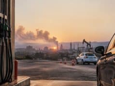 Carburants chers : votre permis pourrait coûter plus cher aux élèves Pompes à essence au premier plan avec une voiture et une raffinerie en arrière-plan sous un coucher de soleil.