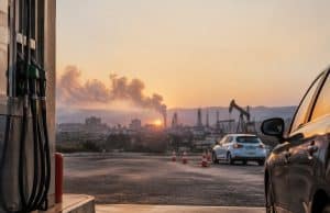 Carburants chers : votre permis pourrait coûter plus cher aux élèves Pompes à essence au premier plan avec une voiture et une raffinerie en arrière-plan sous un coucher de soleil.