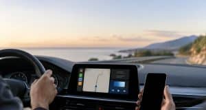Android Auto : Google pourrait exclure les appareils sous Android 9 et 10 Conducteur tenant un smartphone dans sa main droite face au tableau de bord d'une voiture avec un système d'infodivertissement.