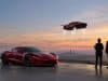 Le nouveau Tesla Roadster face aux régulateurs américains Une Tesla Roadster rouge au sol et une autre volant dans le ciel au coucher du soleil face à deux personnes en costume.