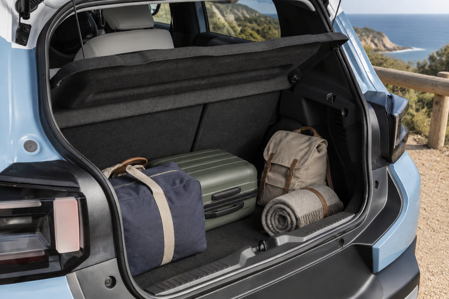 Le coffre ouvert d’une voiture contient une valise, deux sacs et une couverture roulée, avec un paysage côtier en arrière-plan.