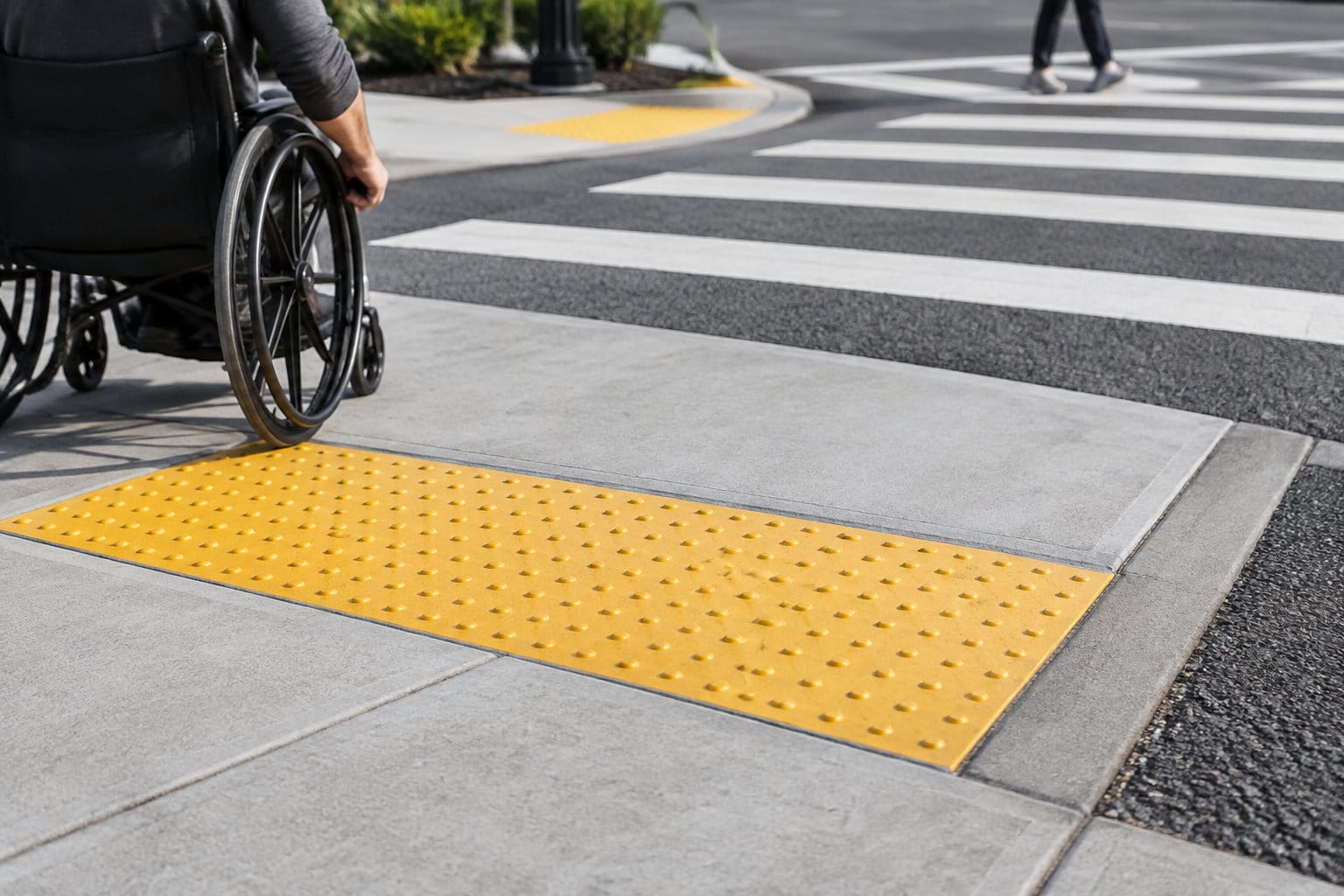 Une personne en fauteuil roulant approche d’un passage piéton, près d’une bande podotactile jaune sur le trottoir.